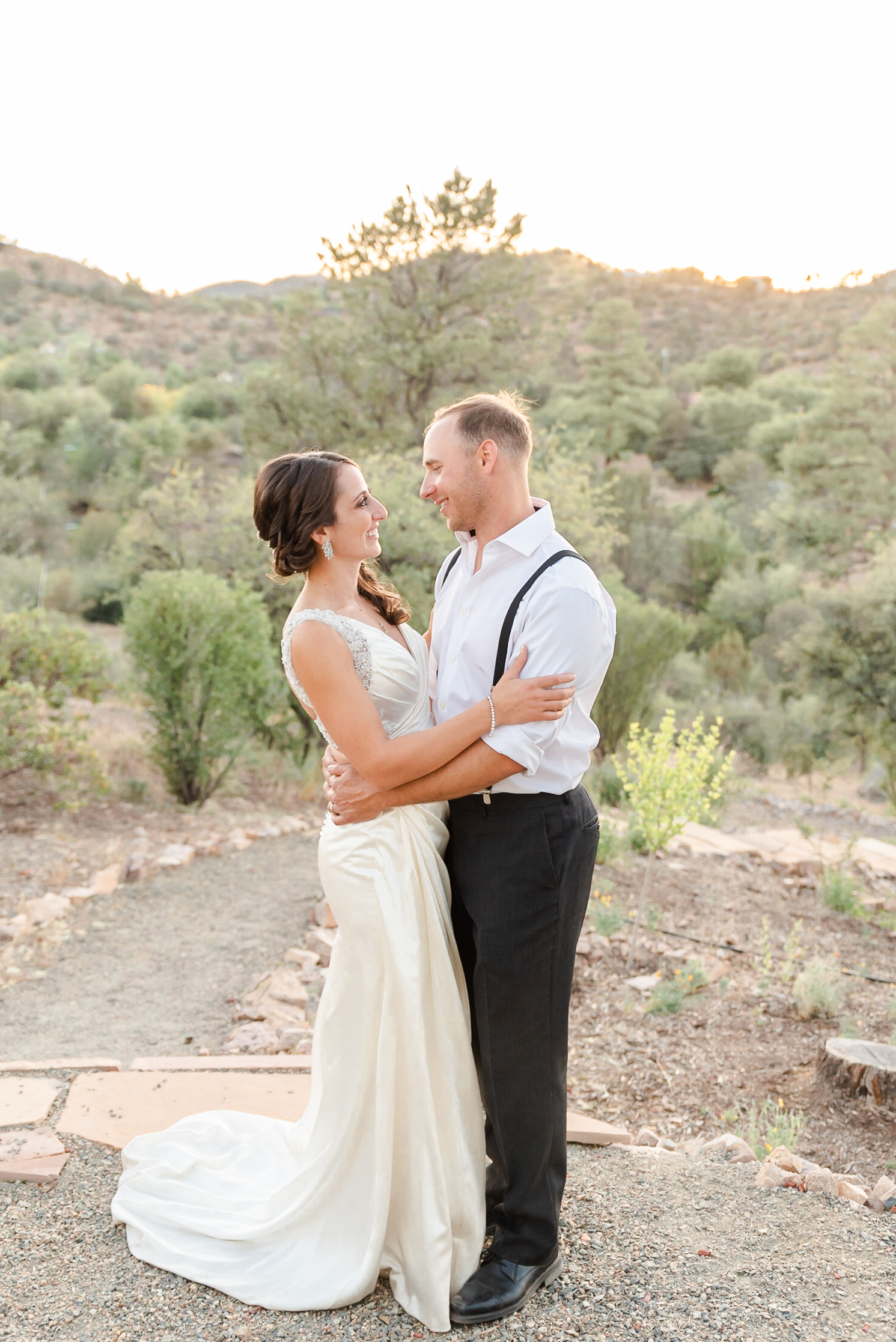 man in suit and woman in white wedding dress embracing each other during sunset on a hill
