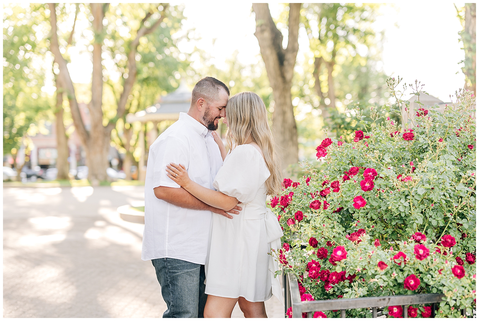 man and wife wearing white standing on green grass in park in front of red roses