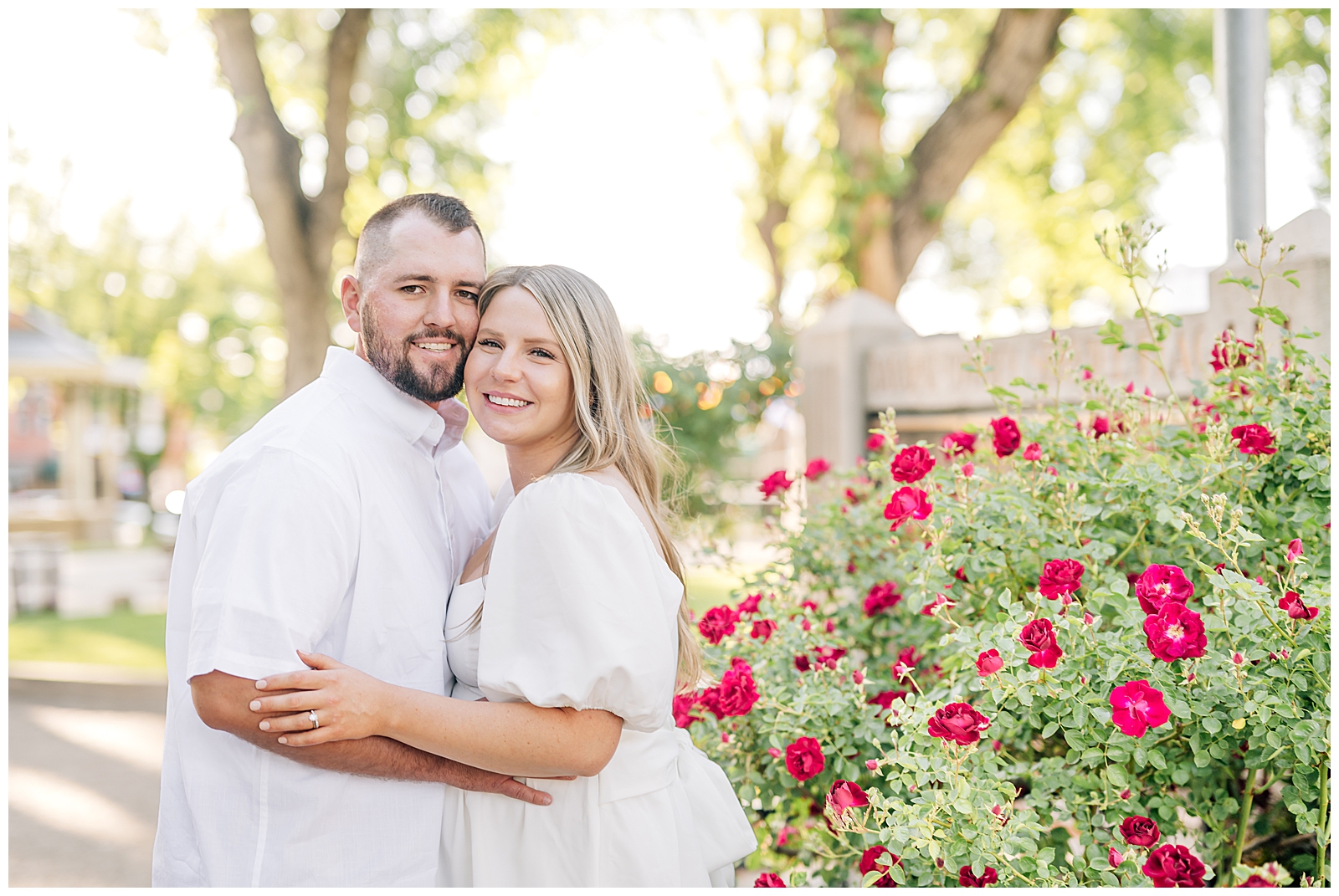 man and wife wearing white standing on green grass in park in front of red roses